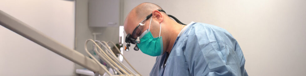 A Brookline dentist wearing magnifying loupes, a mask, and scrubs works on a patient in a modern dental office in Brookline, MA.