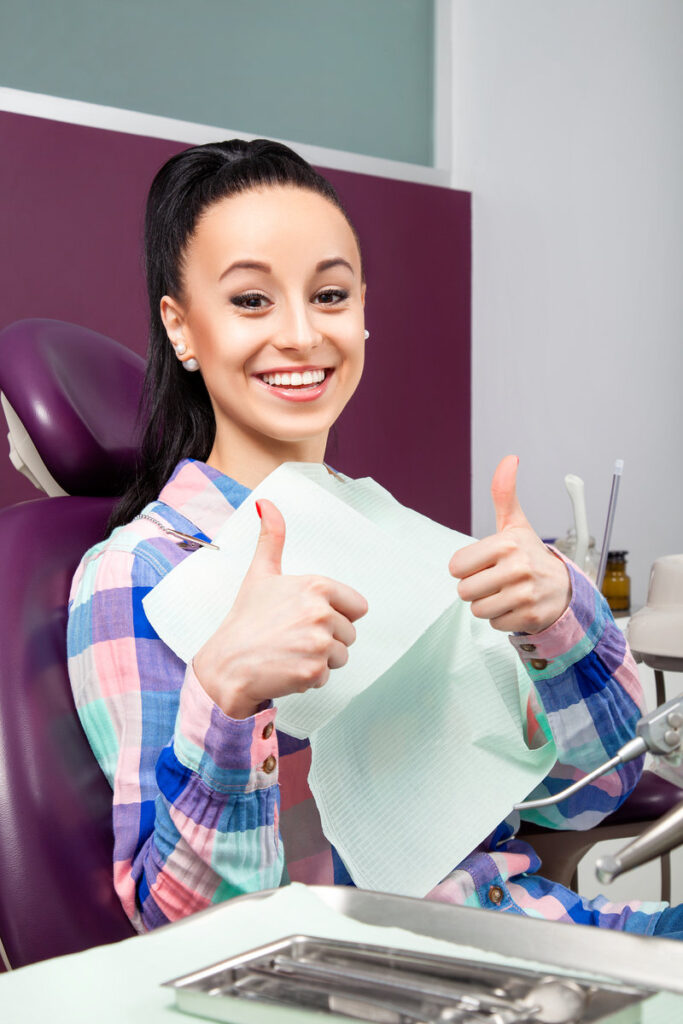 Young woman patient in checkered shirt with perfect straight white teeth with thumbs up waiting for dentist in dental chair and smiling relaxed, ready for a check-up. Beautiful woman smile
