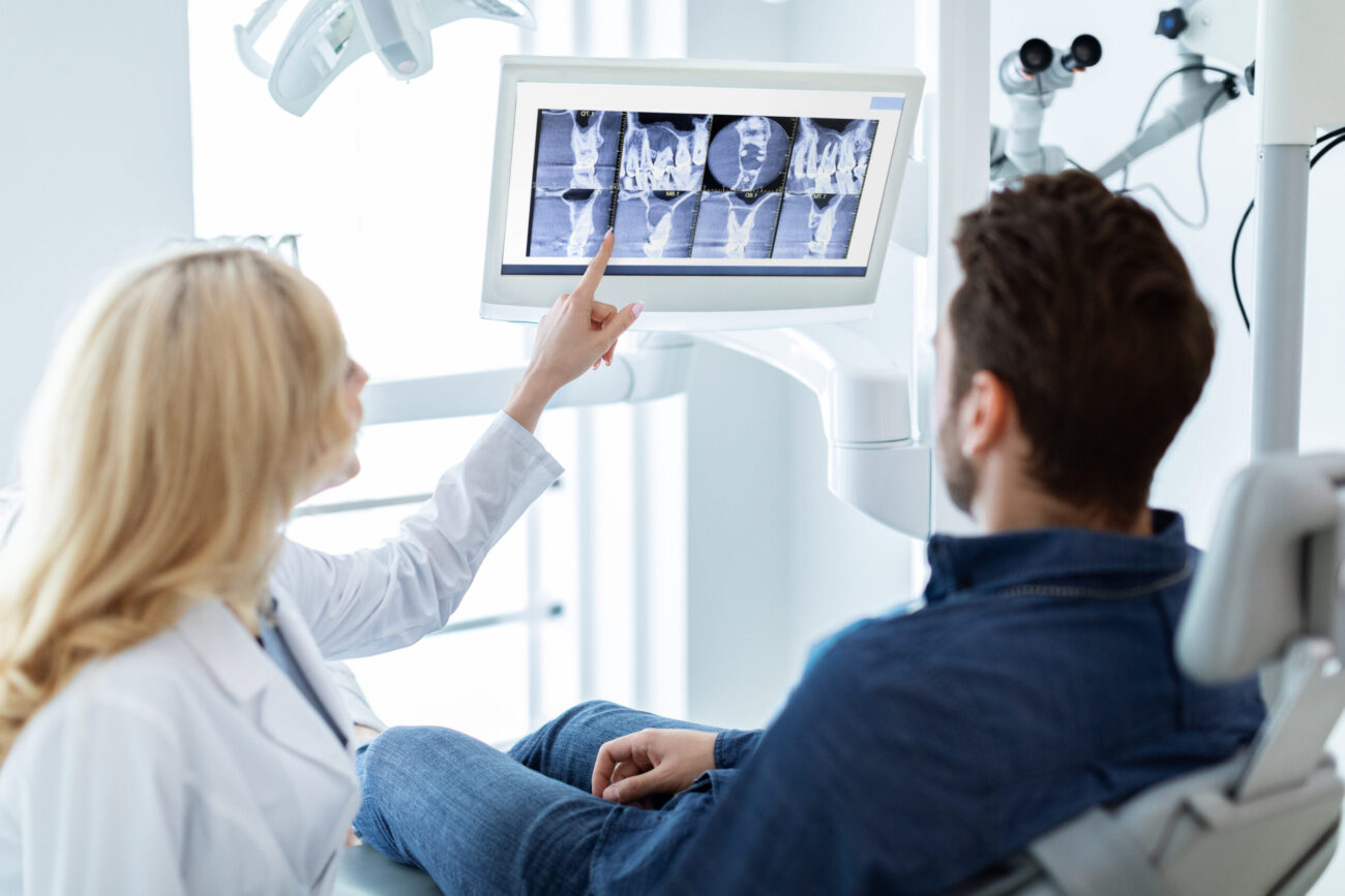 Modern dental clinic concept. Blonde woman dentist having conversation with patient in dental chair, poitning at LED display, showing results of treatment or explaining procedure